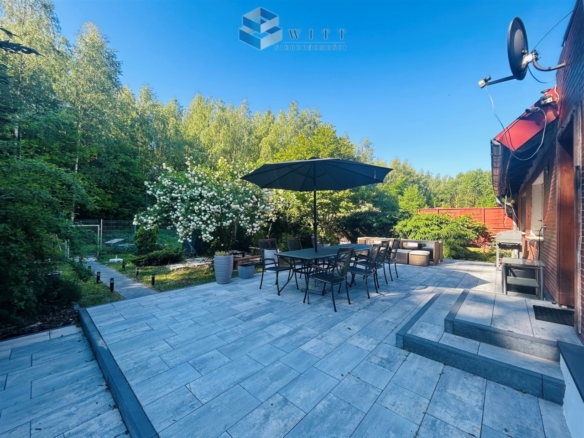 Taras z meblami ogrodowymi i parasolem, otoczony zielenią i kwitnącymi krzewami, w słoneczny dzień.