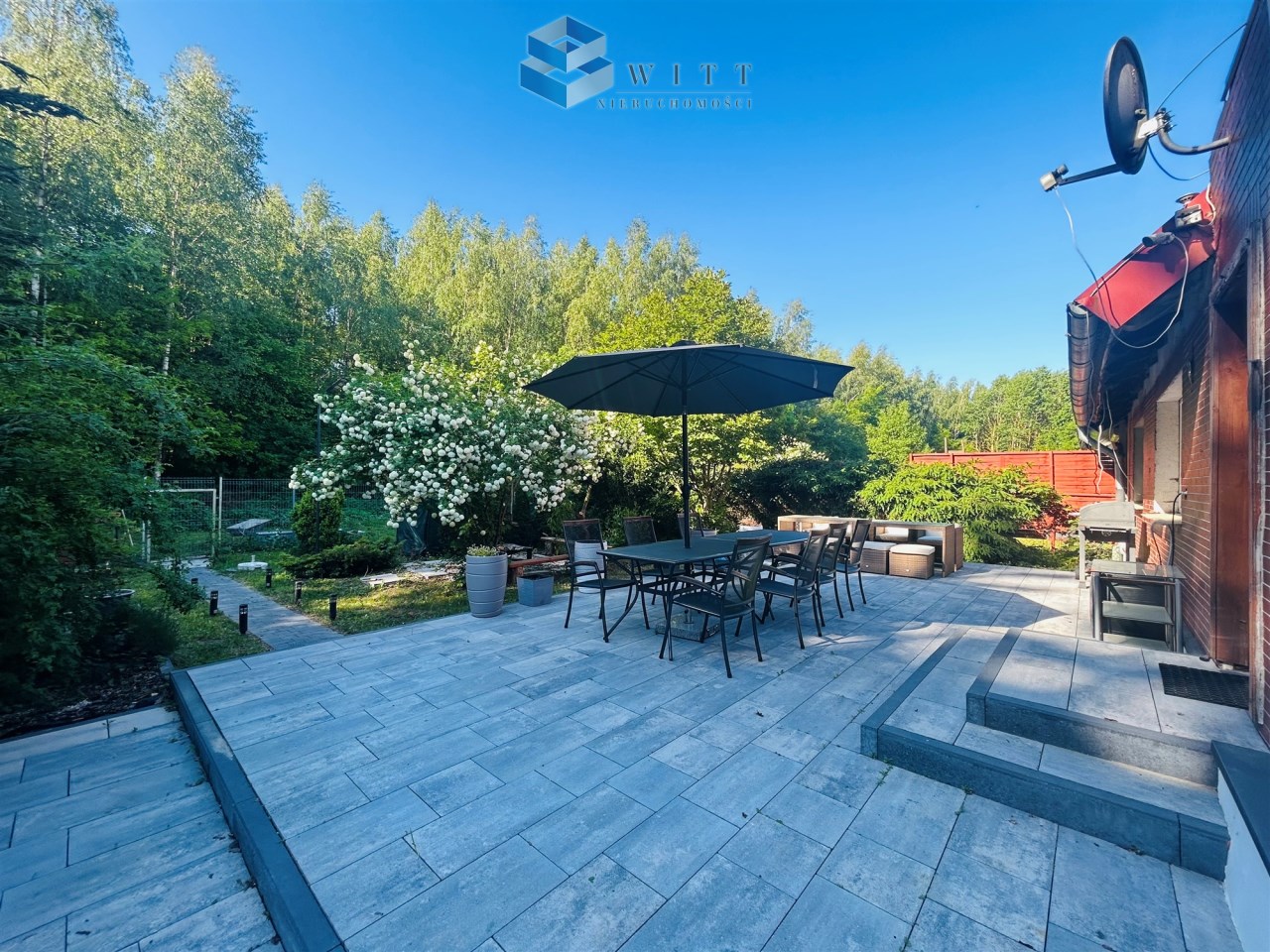 Taras z meblami ogrodowymi i parasolem, otoczony zielenią i kwitnącymi krzewami, w słoneczny dzień.