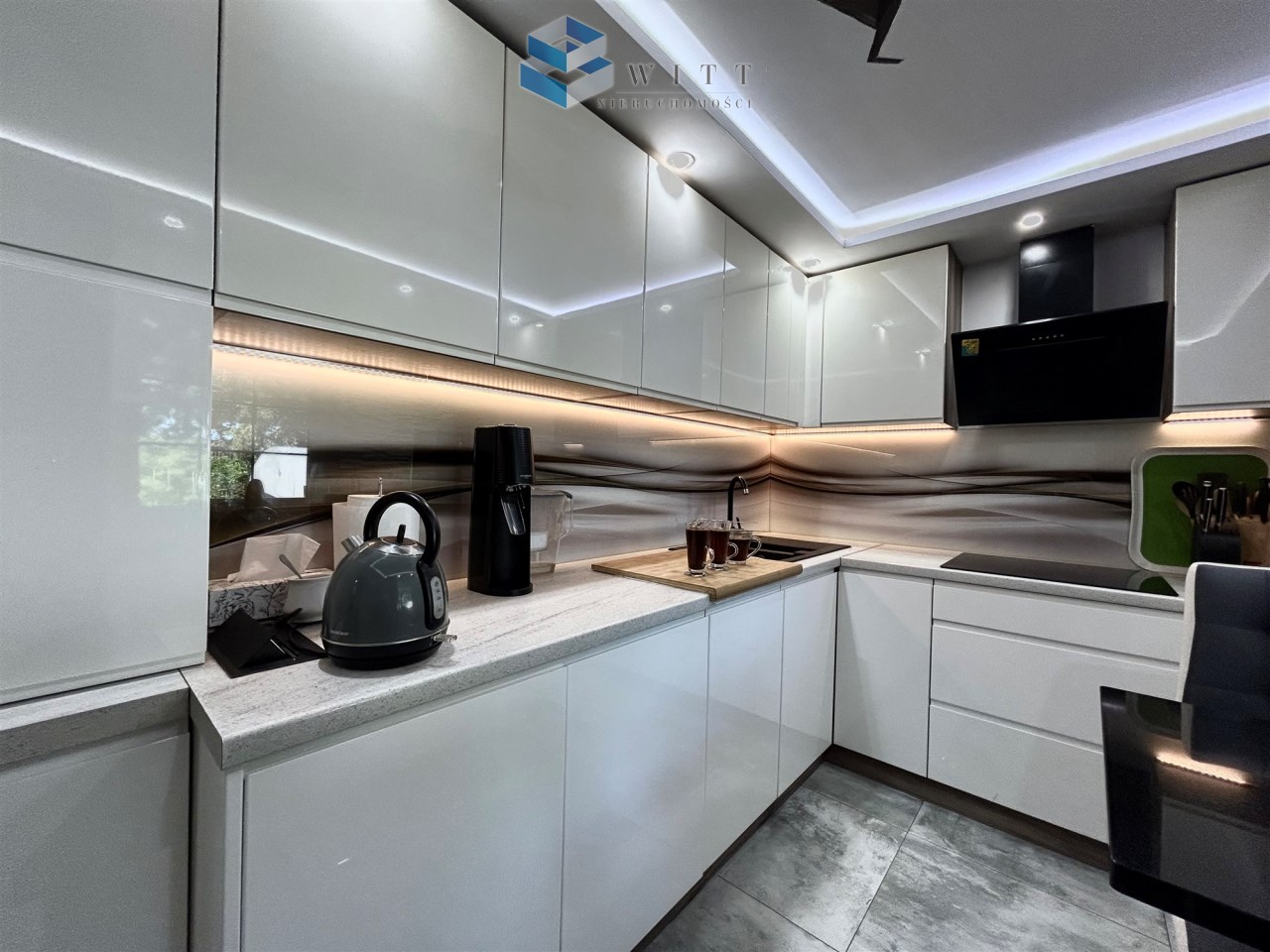 Nowoczesna kuchnia z białymi, błyszczącymi szafkami, szarym czajnikiem, czarnym ekspresem do kawy oraz eleganckim blatem. Oświetlenie LED pod szafkami tworzy przyjemną atmosferę. W tle widoczna jest stylowa ściana z delikatnym wzorem.