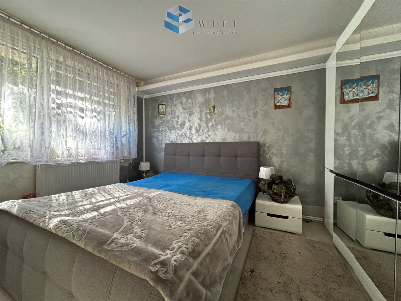 Wnętrze sypialni z dużym łóżkiem, szarym zagłówkiem i niebieską pościelą. Na ścianach dekoracyjne obrazy, a obok łóżka biała szafka nocna z lampką. Duże okno z firankami wpuszcza naturalne światło.