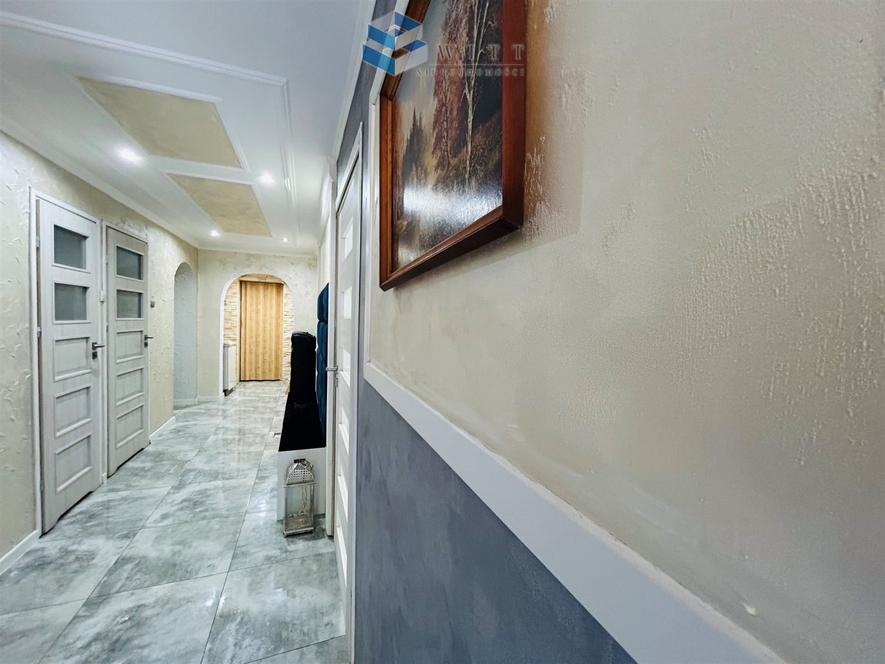 Widok na nowoczesny korytarz z białymi drzwiami, jasnymi ścianami i płytkami podłogowymi w odcieniach szarości. Na ścianie znajduje się obraz w ramie, a w tle widoczna jest łukowata nisza oraz drewniana szafa.