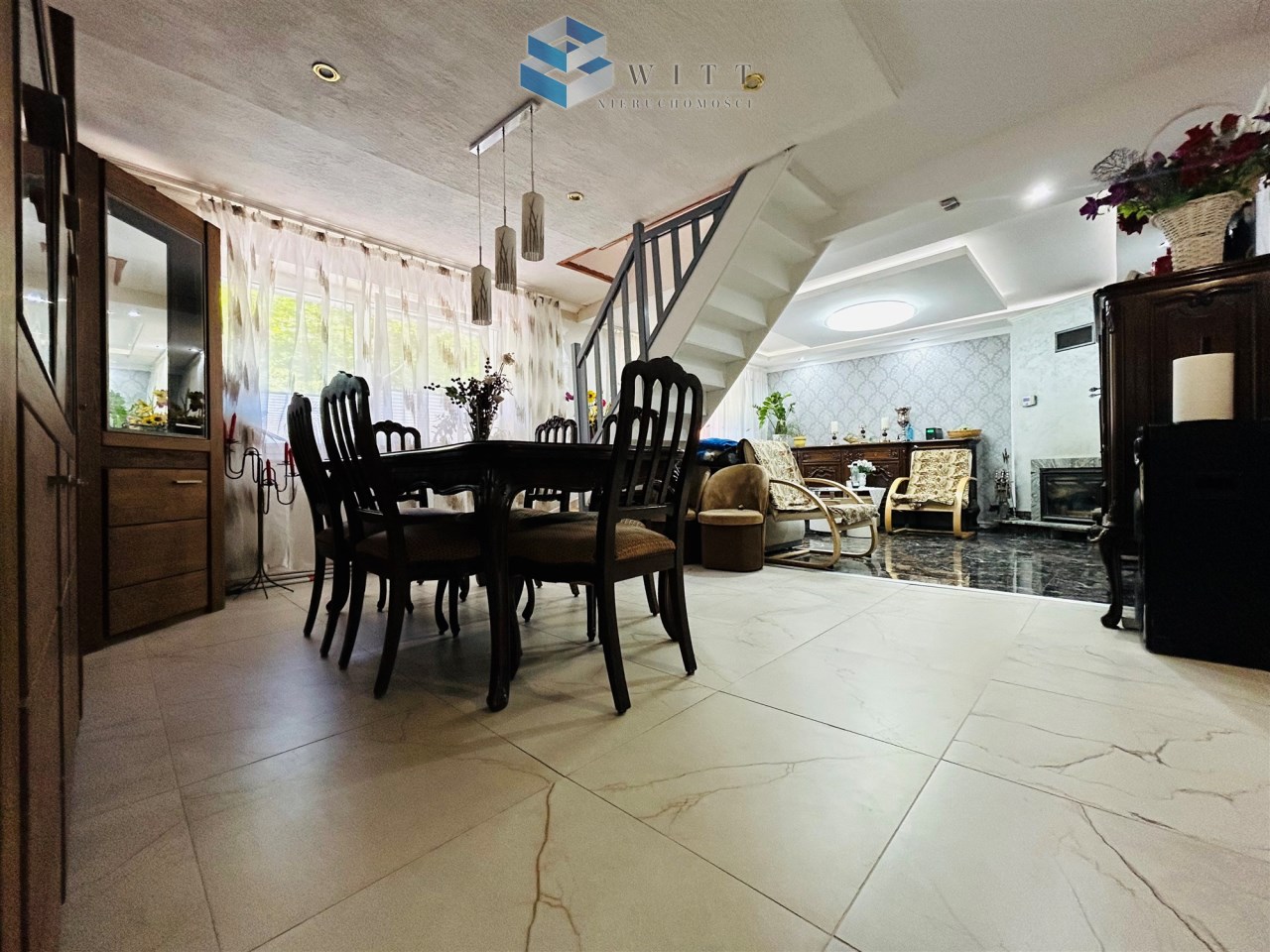 Wnętrze nowoczesnego salonu z jadalnią, drewniany stół z krzesłami, kominek, eleganckie oświetlenie i duże okna.