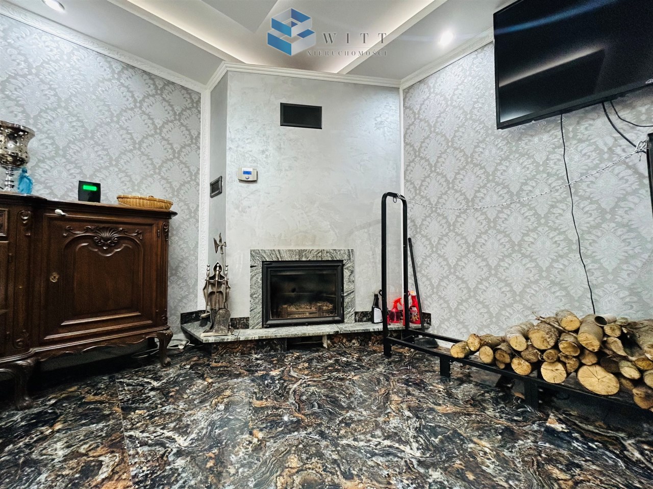 Wnętrze z kominkiem, drewnianą szafą, telewizorem na ścianie oraz podłogą w marmurowym wzorze. Obok kominka stos drewna i akcesoria kominkowe.