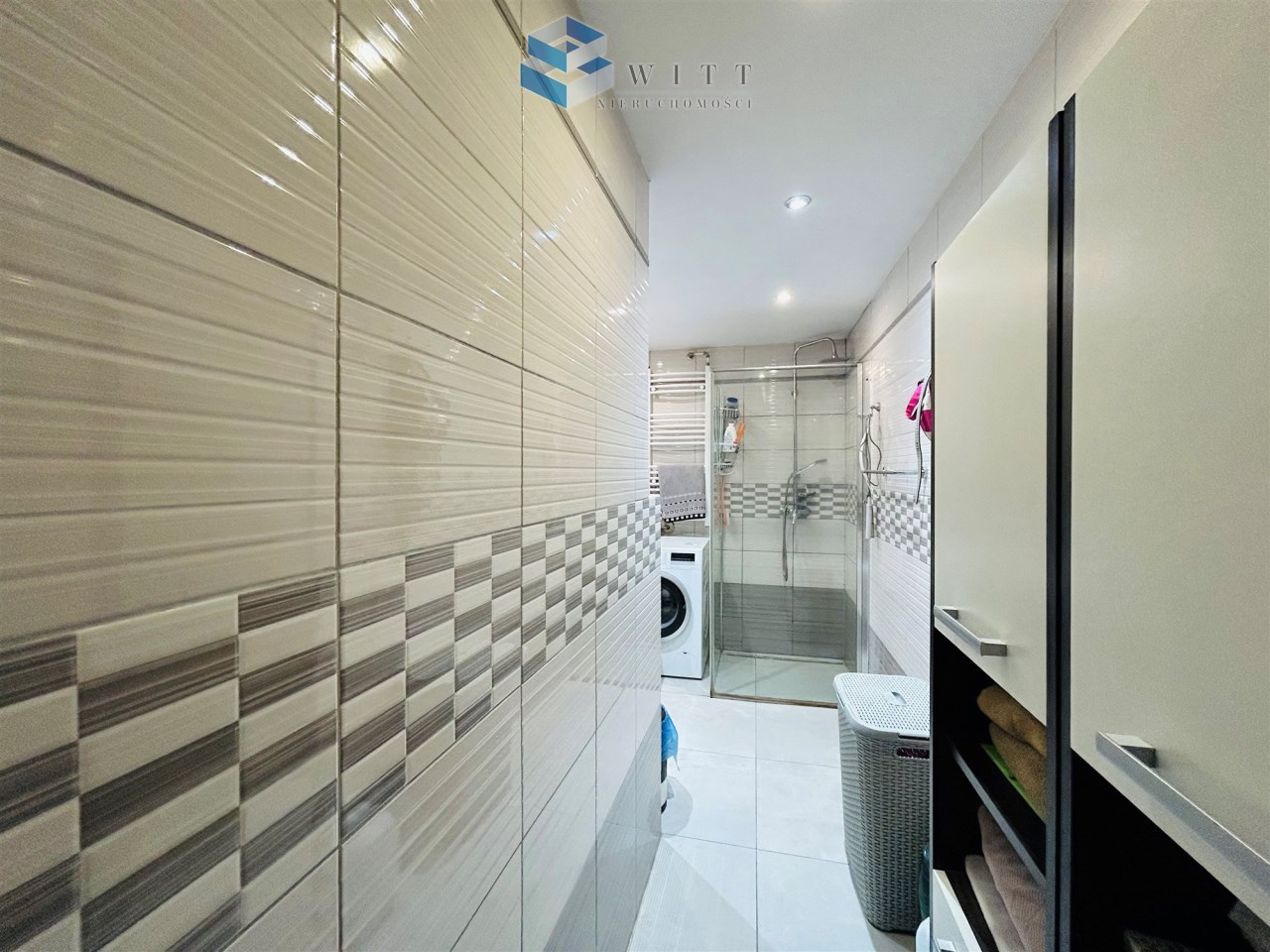 Wnętrze nowoczesnej łazienki z prysznicem, pralką i szafkami na ręczniki. Ściany pokryte jasnymi kafelkami z dekoracyjnym wzorem. Dobrze oświetlone pomieszczenie.