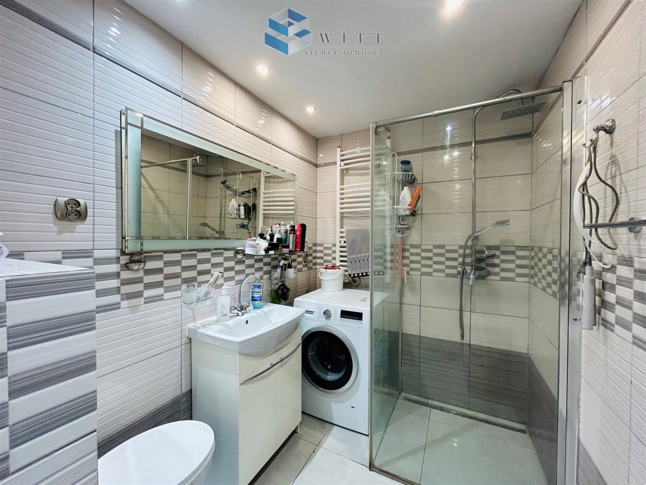 Nowoczesna łazienka z prysznicem, umywalką, pralką i lustrem. Ściany wyłożone jasnymi płytkami w odcieniach szarości. Oświetlenie sufitowe oraz grzejnik drabinkowy.