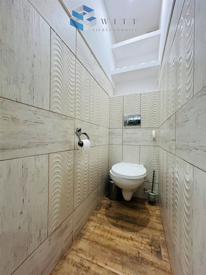 Toaleta z nowoczesnym wykończeniem, jasnymi kafelkami i drewnianą podłogą. W tle widoczna toaleta, uchwyt na papier toaletowy oraz miejsce na akcesoria.