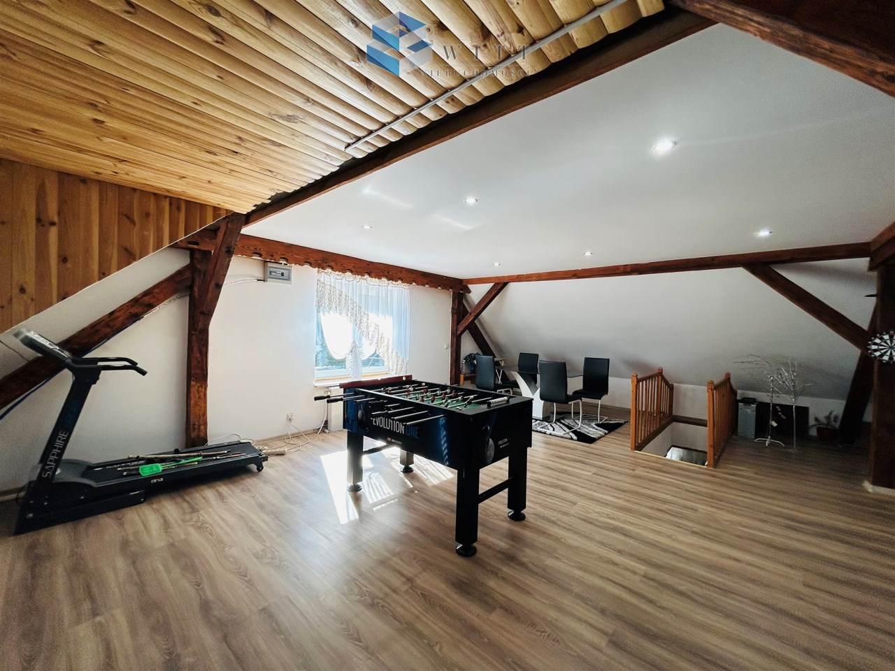 Przestronny poddaszowy pokój z drewnianymi belkami, stołem do piłkarzyków, bieżnią oraz nowoczesnymi krzesłami. Naturalne światło wpada przez okno, oświetlając jasną podłogę.