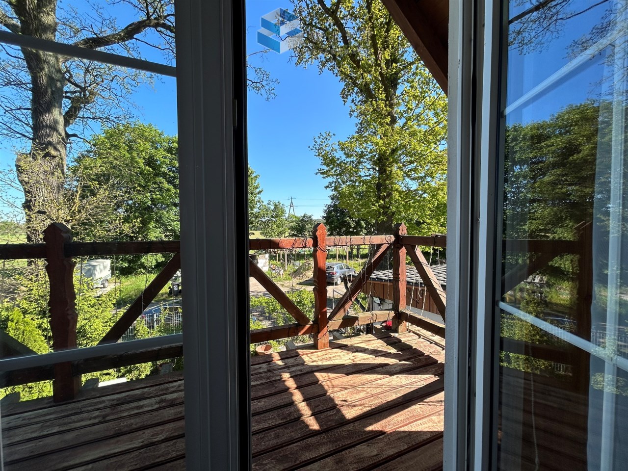 Widok z okna na drewniany taras z balustradą, otoczony zielenią drzew i błękitnym niebem.