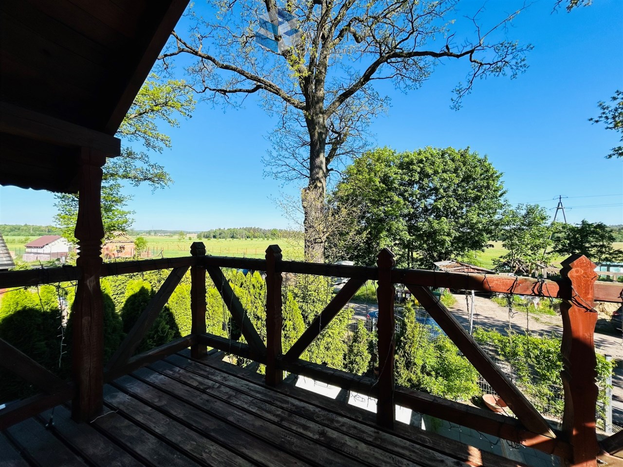 Widok z drewnianego tarasu na zielony krajobraz, drzewa i niebieskie niebo. W tle widać domki oraz pola.
