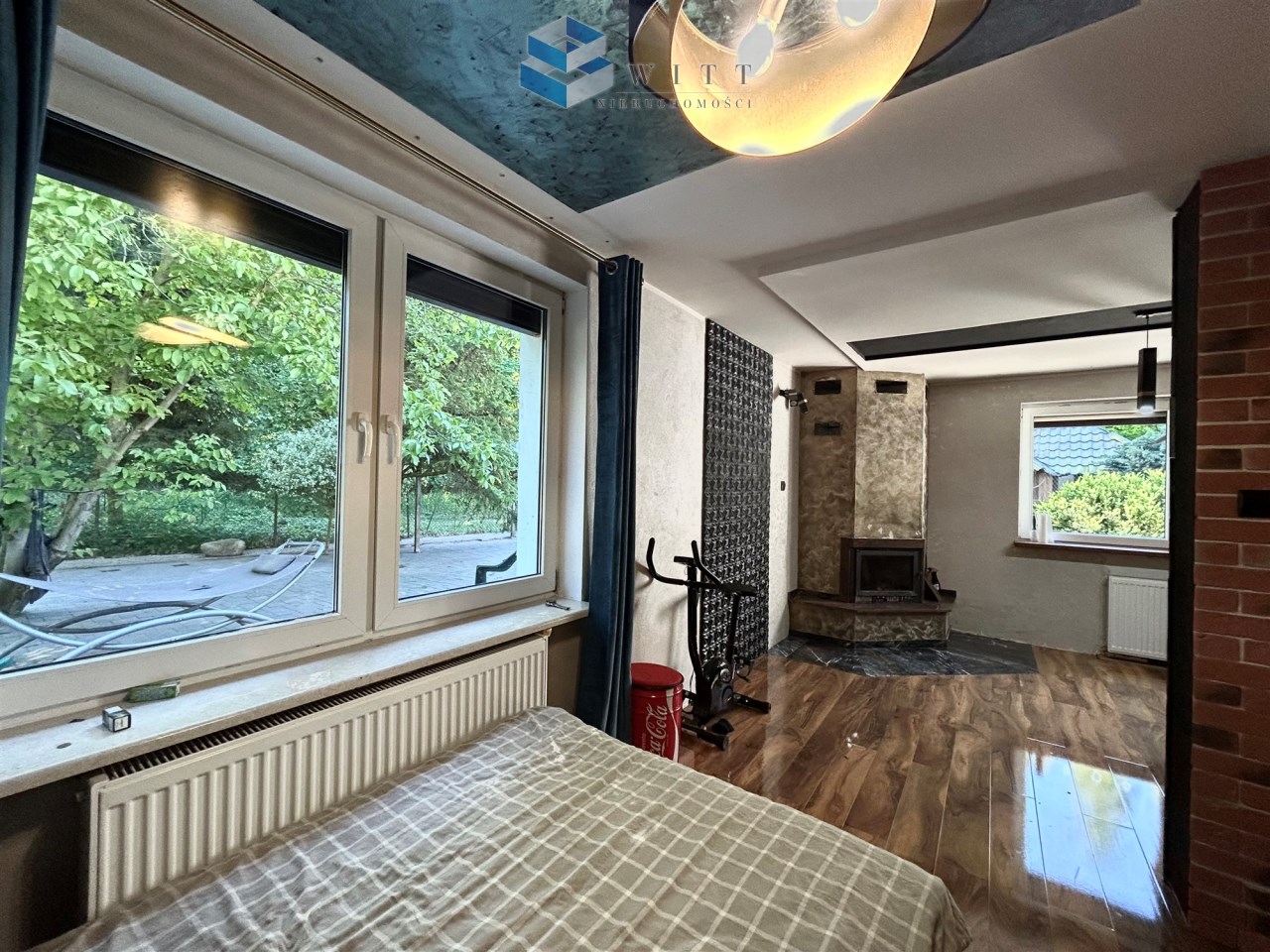 Widok wnętrza nowoczesnego pomieszczenia z oknami wychodzącymi na zewnątrz, z drewnianą podłogą, kominkiem i elementami dekoracyjnymi. W tle widać zieleń ogrodu i sprzęt fitness.