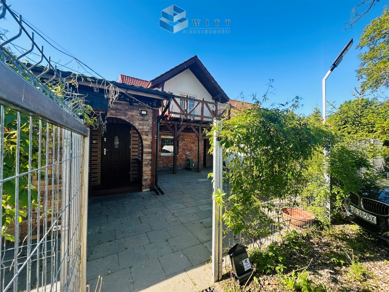 Widok na dom z ogrodzeniem, z zaroślami i podjazdem. Na pierwszym planie widoczna brama i architektura budynku z drewnianymi elementami. Jasne niebo w tle.