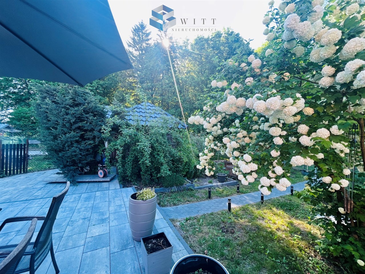 Widok na przytulny ogród z kwitnącymi krzewami, kostką brukową i meblami ogrodowymi, z domem w tle.