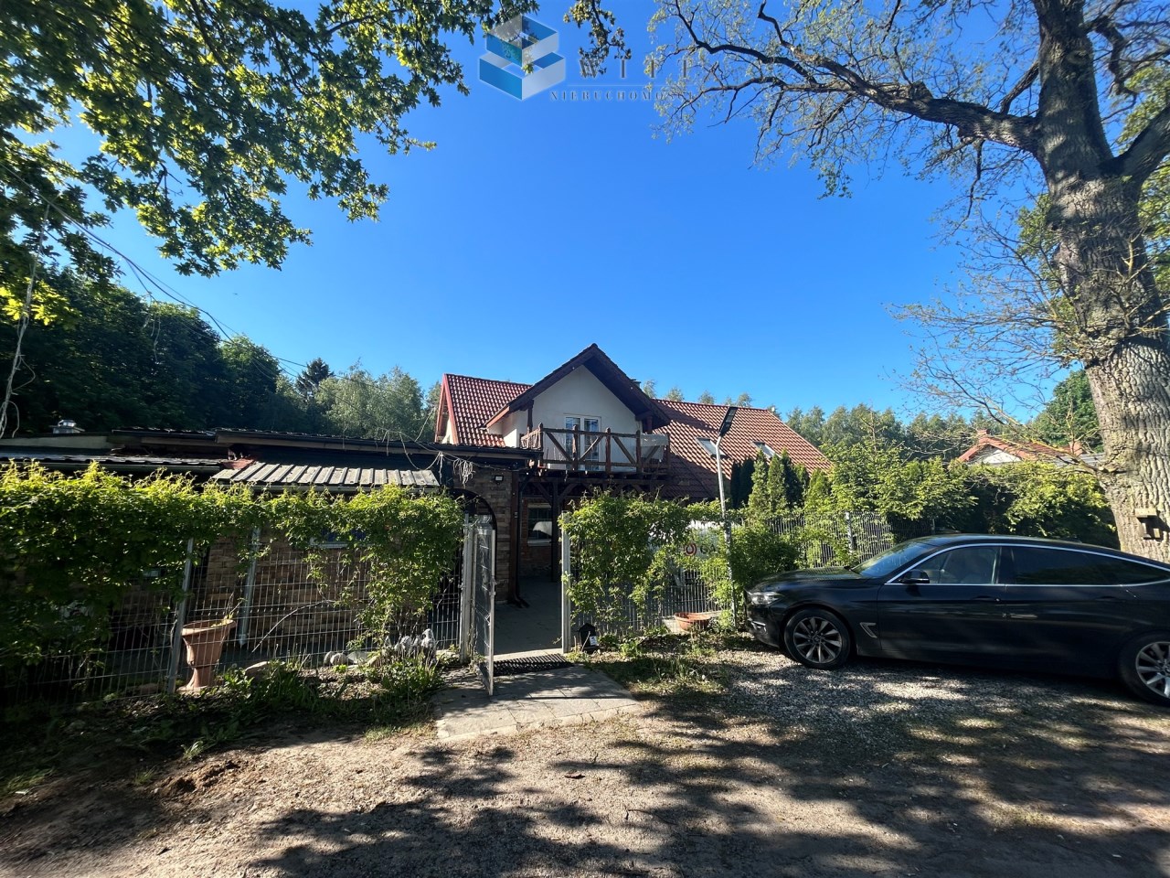 Widok na dom z ogrodem, otoczony zielenią, z czerwonym dachem oraz czarnym samochodem zaparkowanym na podjeździe. Na pierwszym planie widoczne ogrodzenie i drzewa w tle.