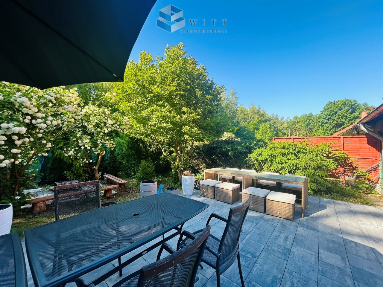 Widok na przytulny ogród z meblami ogrodowymi, stołem i krzesłami, otoczony zielenią i kwitnącymi krzewami. Słoneczne niebo w tle.