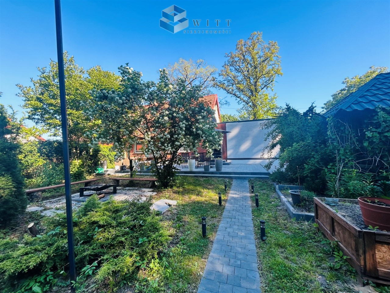 Zielony ogród z drewnianą ławką, kwitnącym drzewem i ścieżką z kostki brukowej prowadzącą do domu. W tle widoczny taras z meblami ogrodowymi oraz zadbane rośliny w donicach. Czyste niebo i słoneczna pogoda.