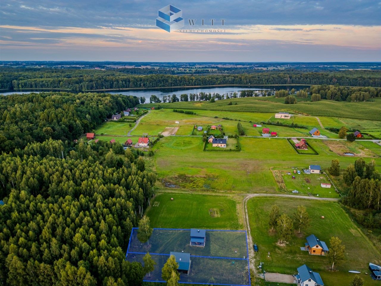 Widok z lotu ptaka na malowniczą wieś z domami otoczonymi zielenią, lasami i rzeką w tle.