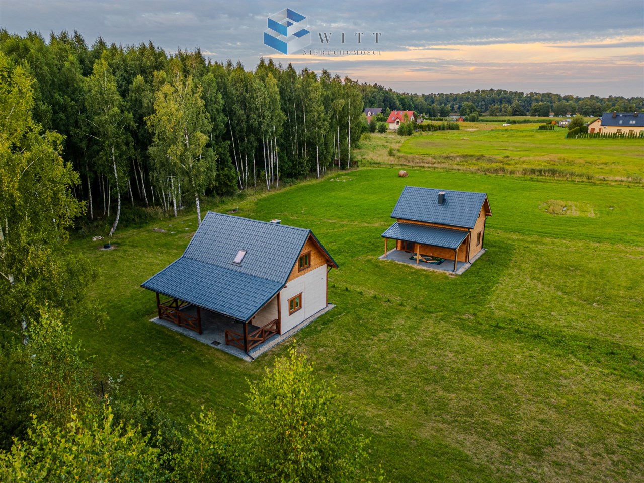 Dwa drewniane domy z dachami pokrytymi blachą, otoczone zielonym trawnikiem i lasem, w spokojnej okolicy.
