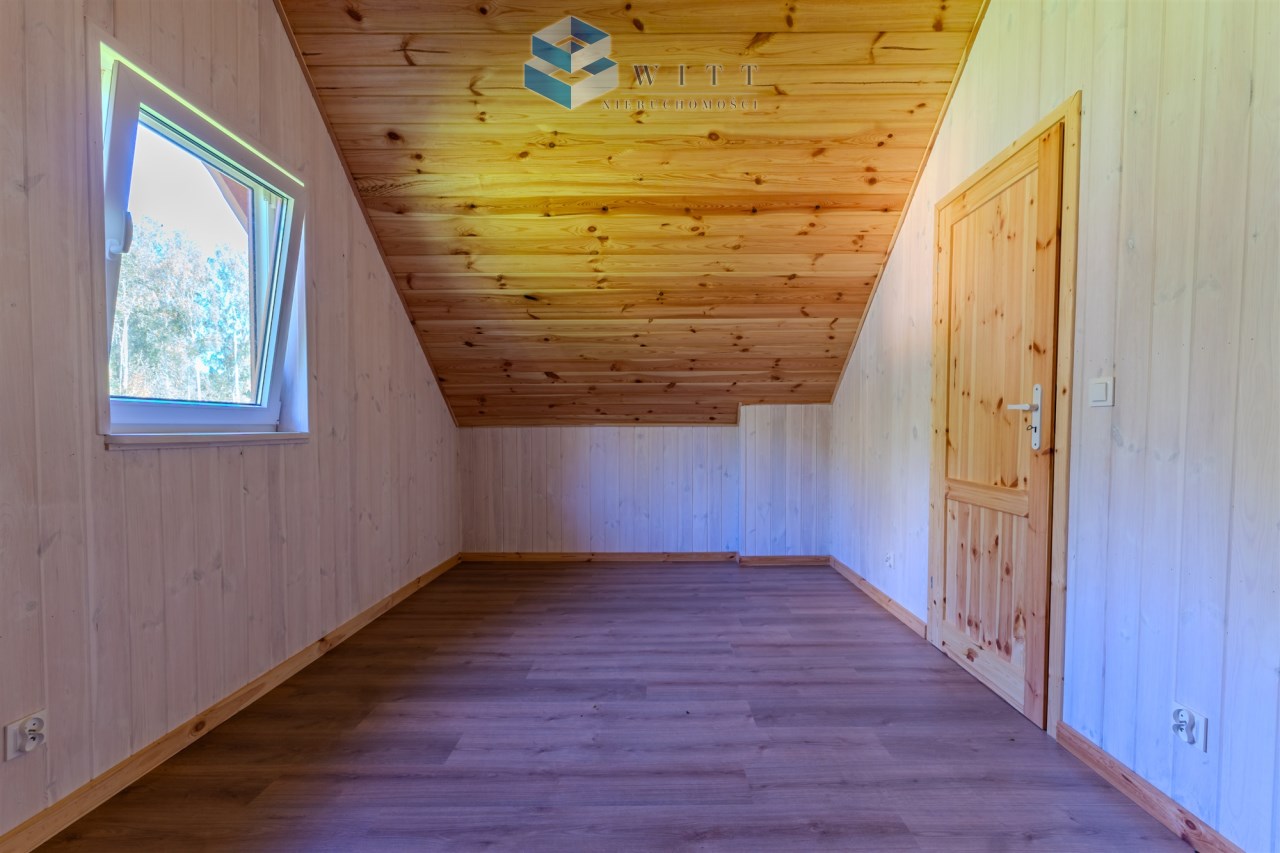 Wnętrze poddasza z drewnianym stropem, jasnymi ścianami i dużym oknem, podłoga z paneli.