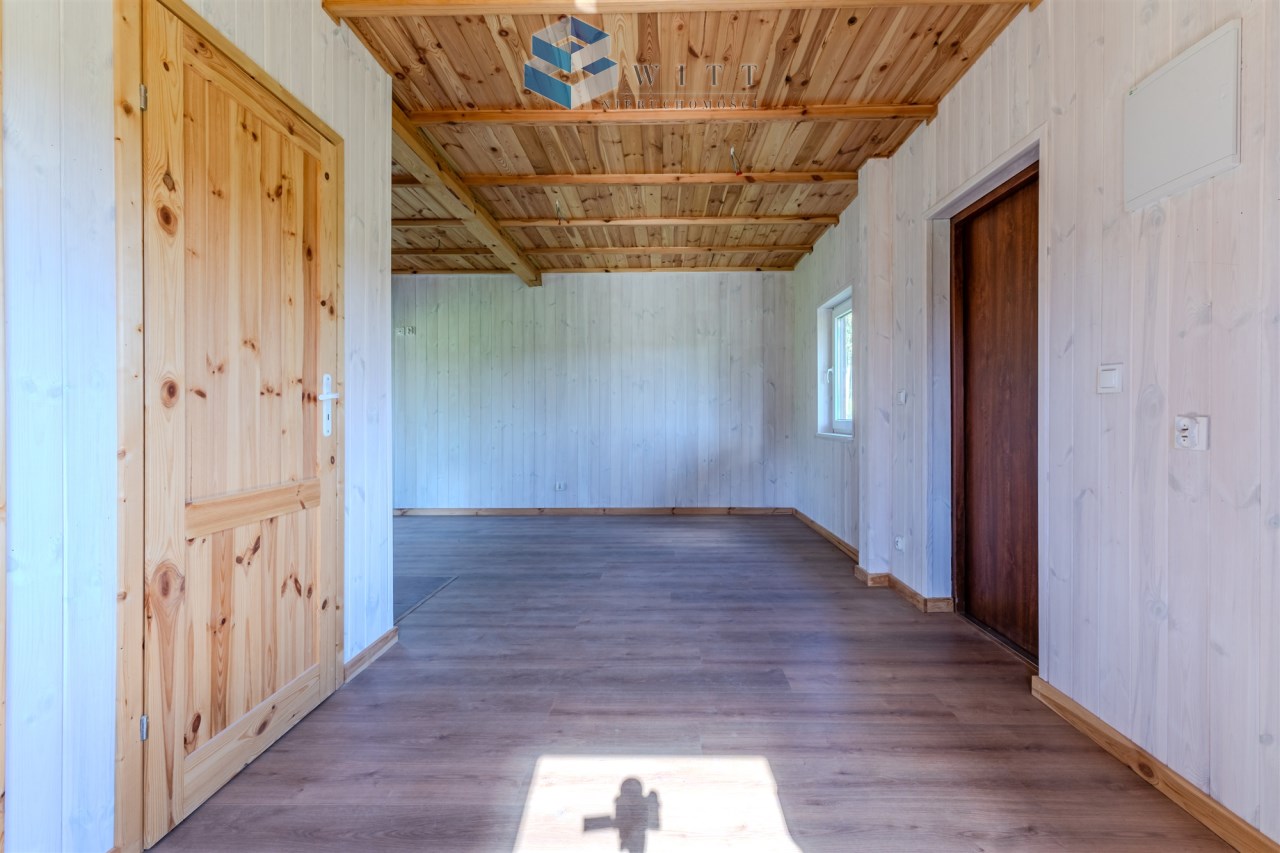 Wnętrze drewnianego pomieszczenia z jasnymi ścianami, drewnianym sufitem i podłogą, z widocznymi drzwiami oraz oknem.