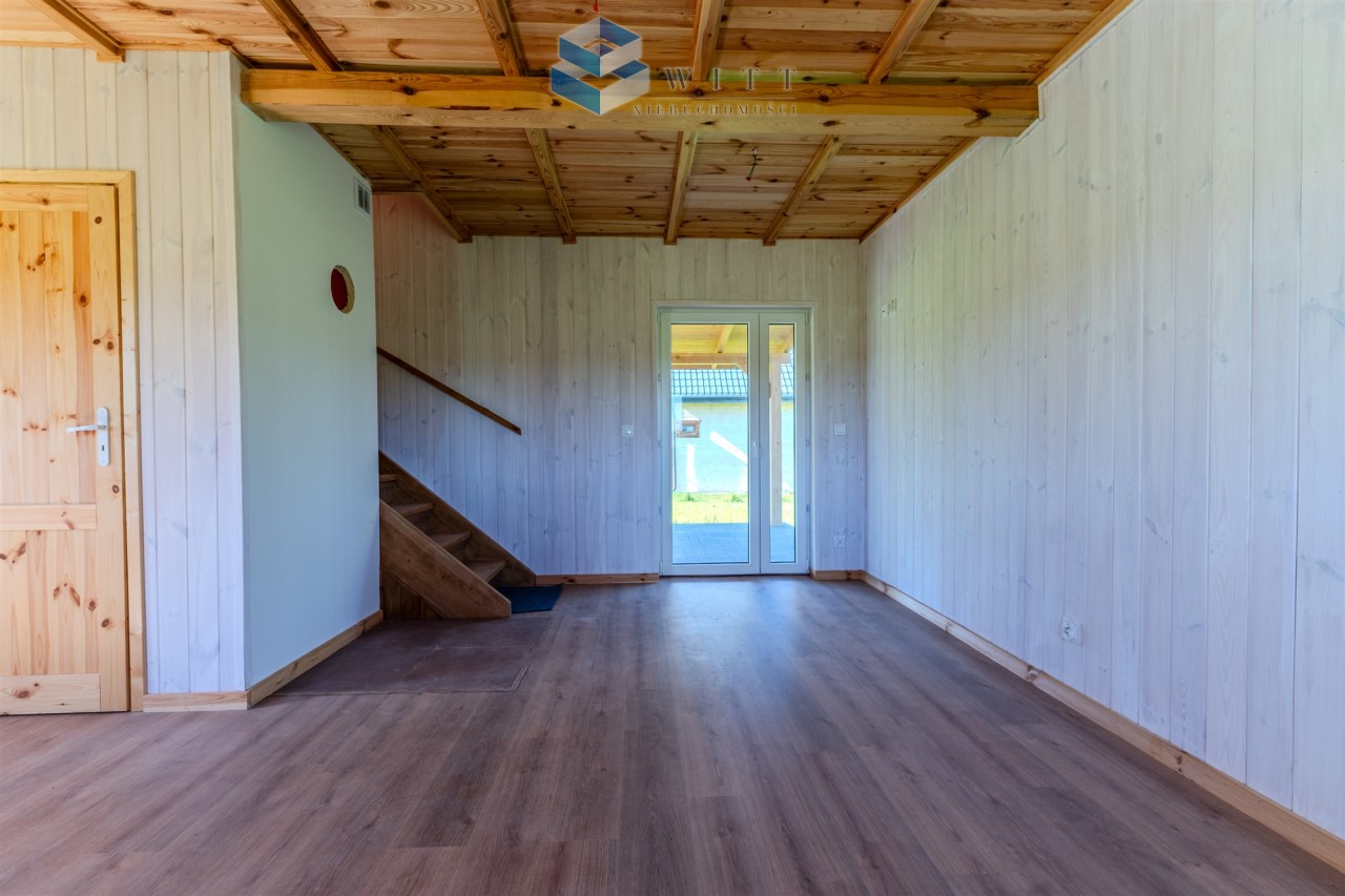 Wnętrze nowoczesnego domu z drewnianymi elementami, jasnymi ścianami i dużymi oknami, prowadzącymi na zewnątrz. Widok na schody oraz drzwi wejściowe.