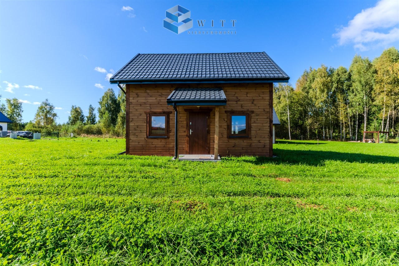 Nowoczesny dom drewniany z czarnym dachem, otoczony zielonym trawnikiem i drzewami w tle, w słoneczny dzień.