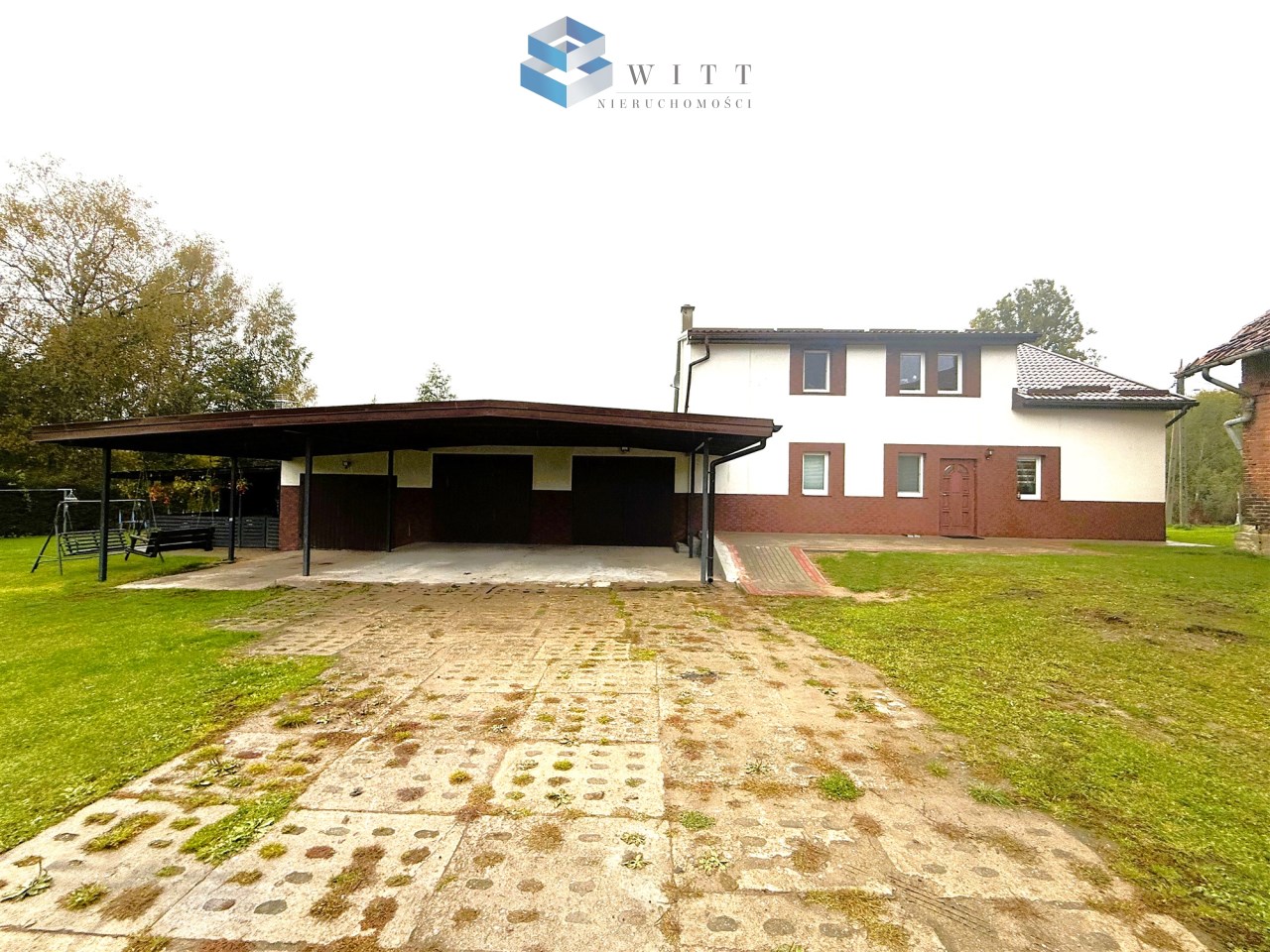 Widok na dom jednorodzinny z przyległym zadaszonym garażem oraz przestronnym podjazdem, otoczony zielenią.