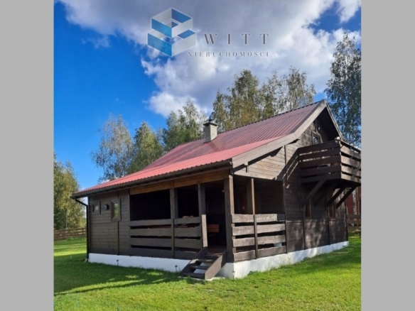 Drewniany domek z czerwonym dachem, otoczony zielonym trawnikiem i drzewami, z zadaszonym tarasem i balkonem.