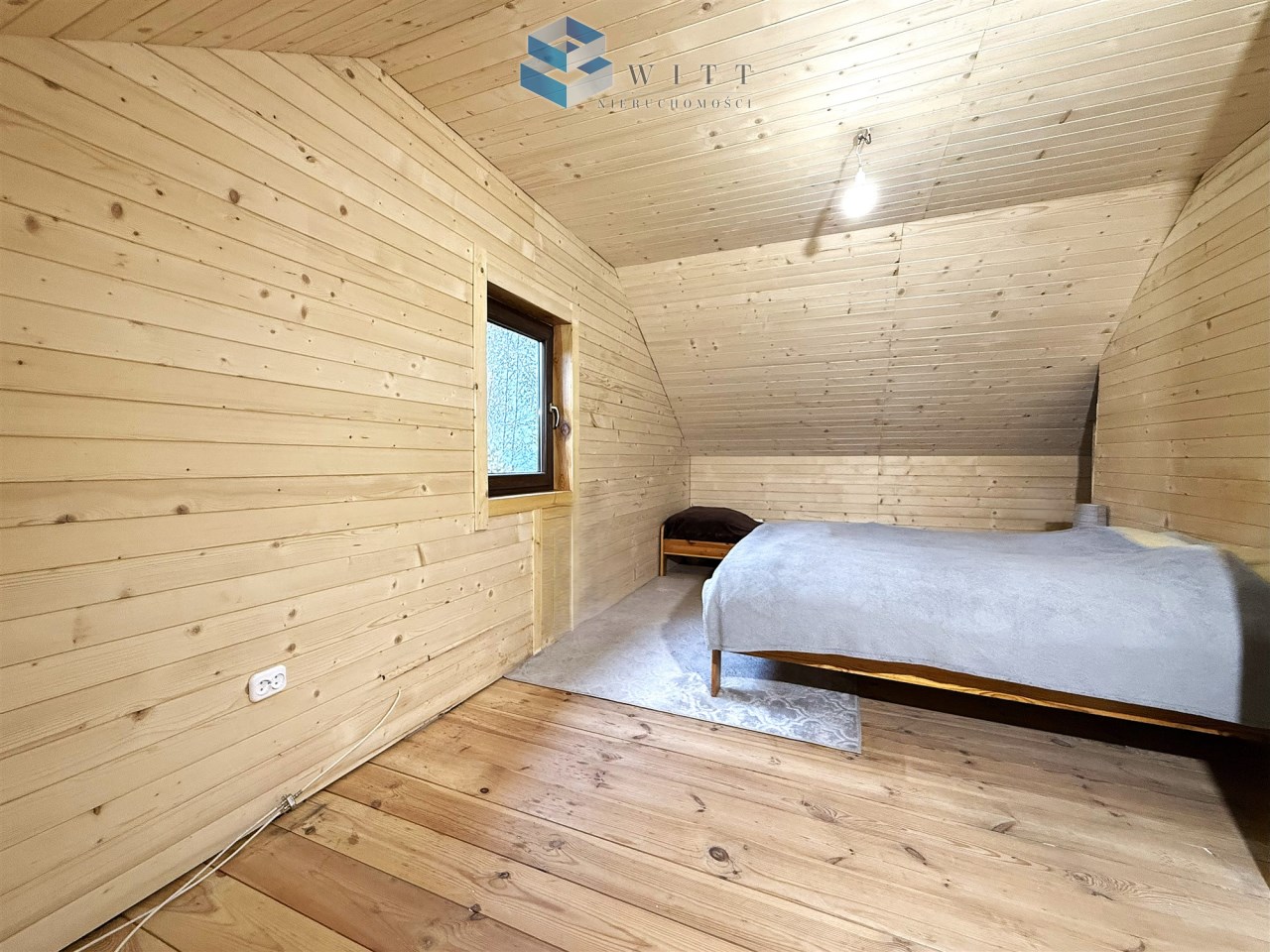 Przytulna sypialnia na poddaszu z drewnianymi ścianami, oknem i łóżkiem z szarym narzutem.