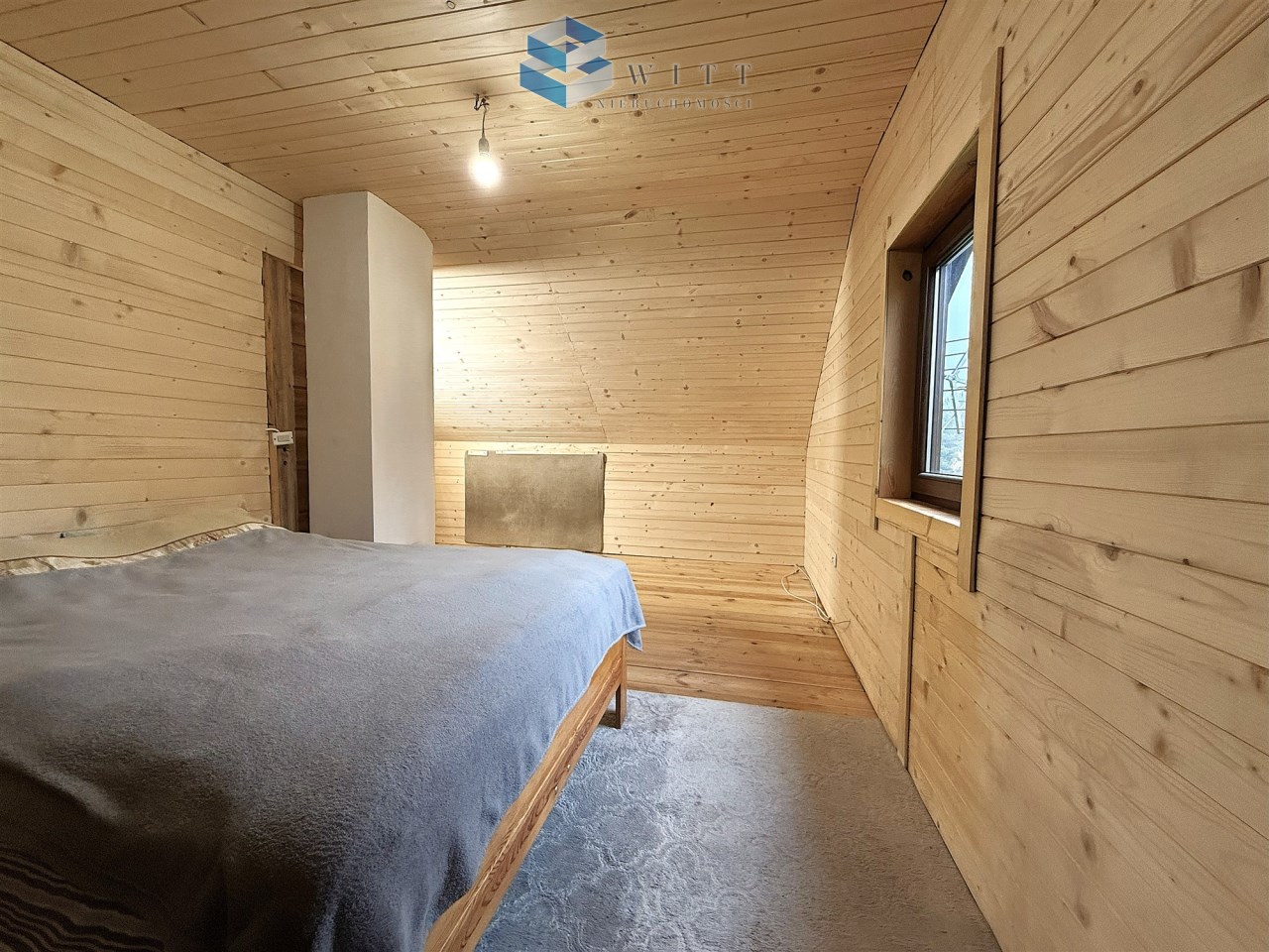 Wnętrze drewnianej sypialni z podwójnym łóżkiem, jasnymi ścianami i oknem, które zapewnia naturalne światło.