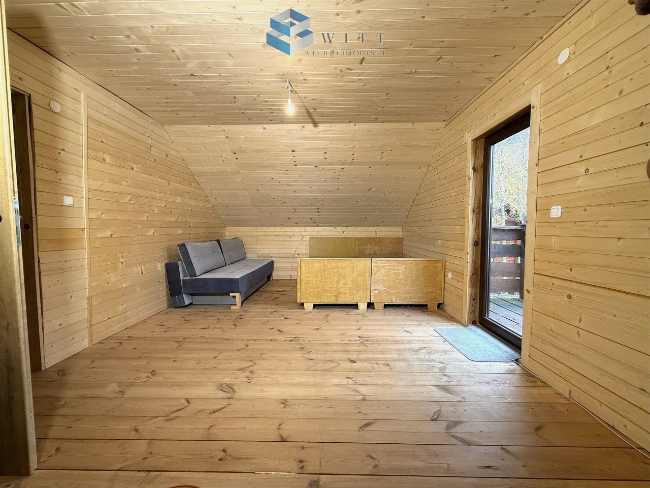 Wnętrze drewnianego pomieszczenia z szarym sofą, drewnianą szafką i dużymi oknami, które prowadzą na balkon.