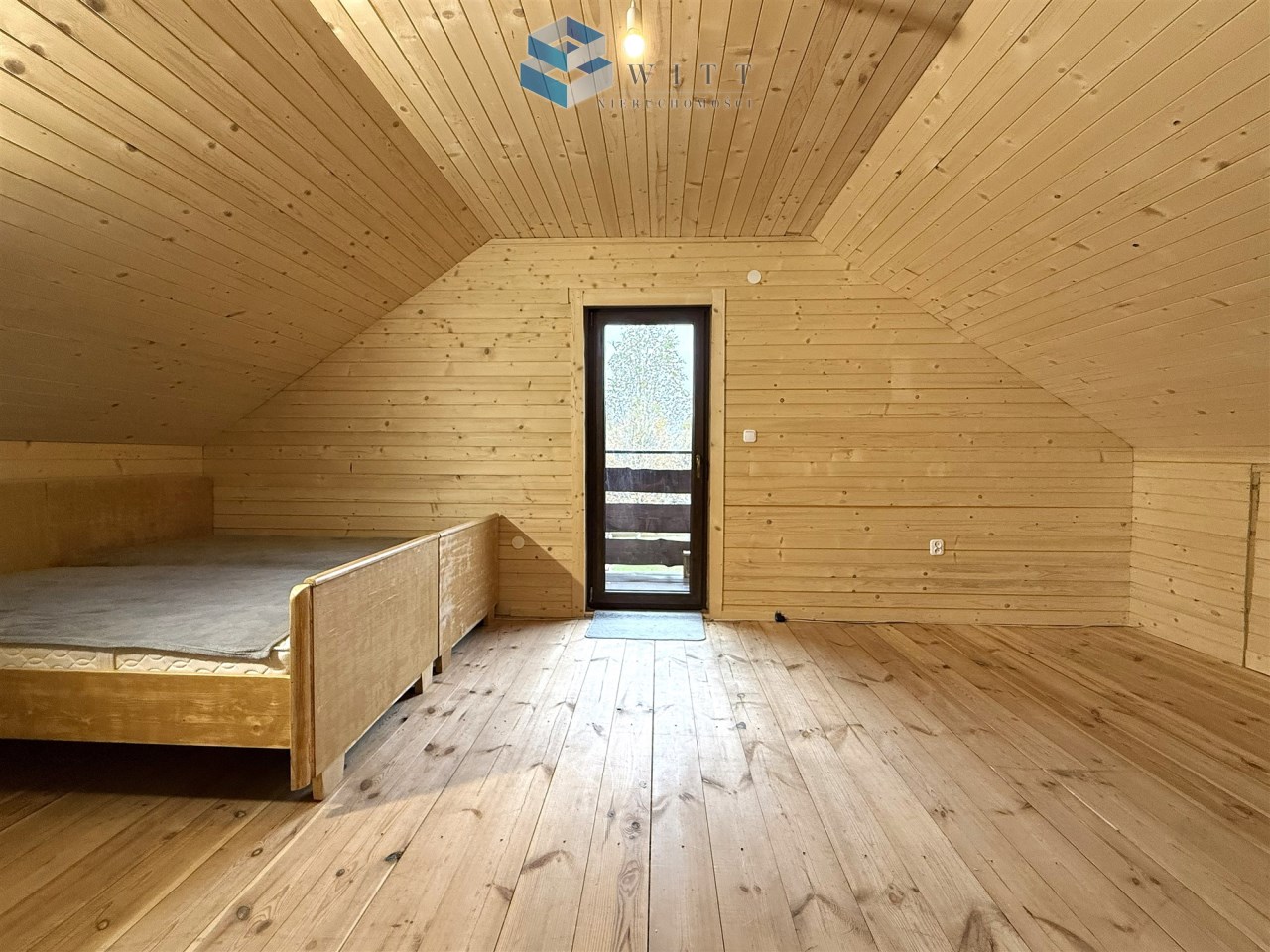 Pusty pokój na poddaszu z drewnianymi ścianami i podłogą, z dużym oknem balkonowym. Wnętrze jasne i przestronne, z łóżkiem po lewej stronie.