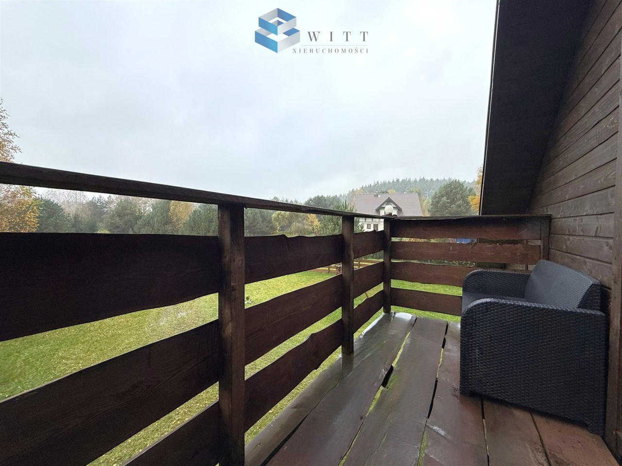 Widok na balkon z drewnianym ogrodzeniem, z meblem wypoczynkowym, otoczony zielenią i drzewami w tle, w pochmurny dzień.