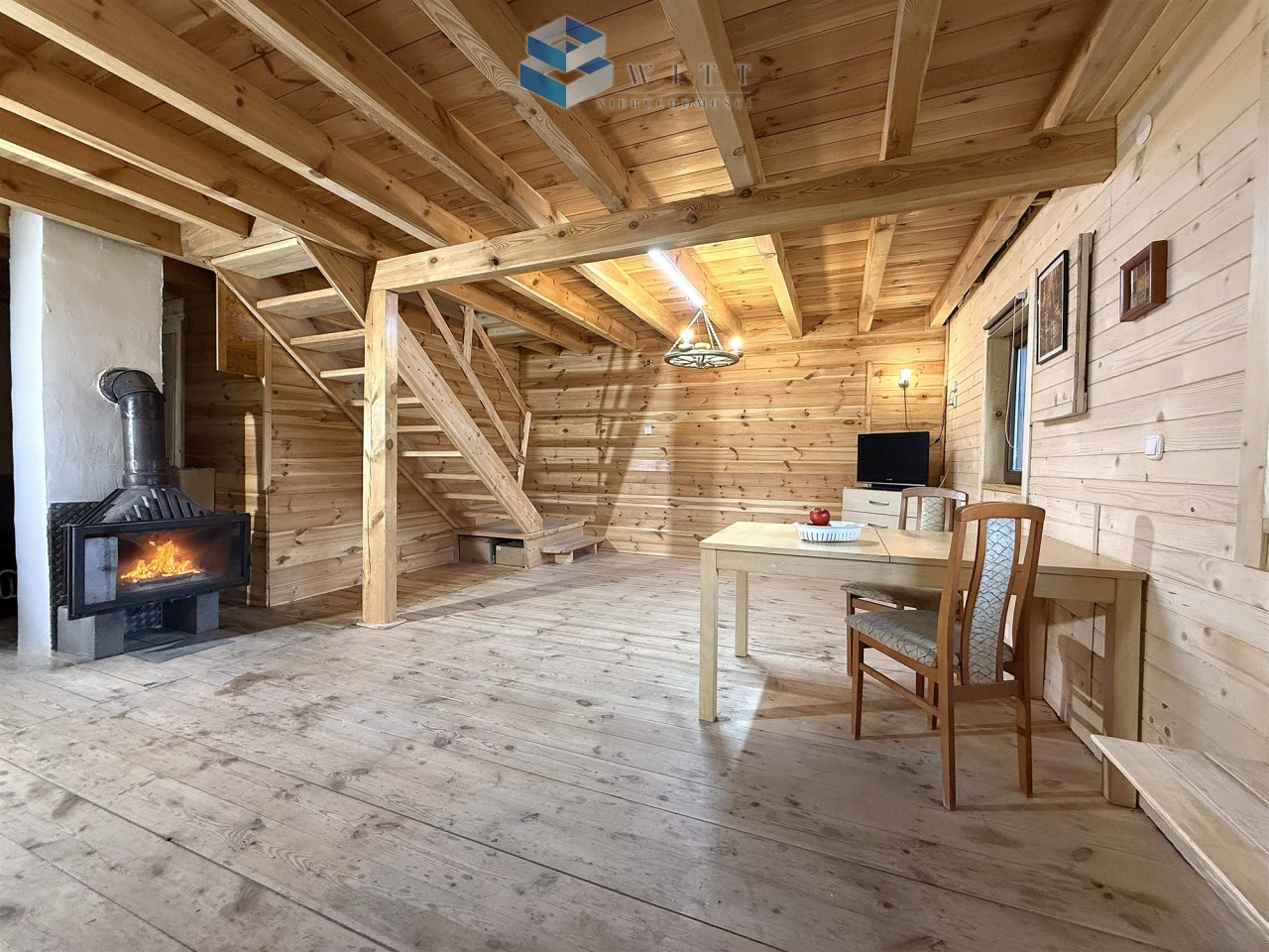 Wnętrze drewnianego domku z kominkiem, drewnianymi schodami i stołem z krzesłem. Naturalne drewno na ścianach i podłodze, przytulna atmosfera.