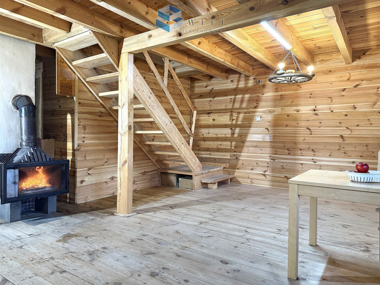 Wnętrze drewnianego domku z kominkiem, schodami i jasnym oświetleniem. Na stole znajduje się jabłko, a ściany pokryte są drewnianymi panelami.