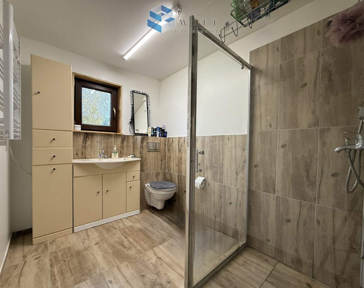 Nowoczesna łazienka z prysznicem, umywalką i toaletą, wykończona w jasnych kolorach, z drewnopodobnymi płytkami na podłodze i ścianach. W tle okno zapewniające naturalne światło.