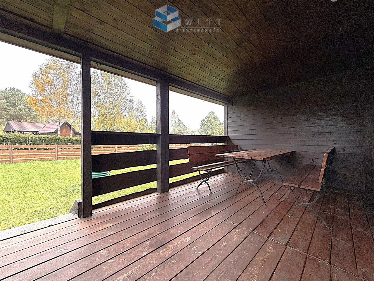 Zadaszony taras z drewnianą podłogą i meblami ogrodowymi, widok na zielony trawnik i drzewa w tle.