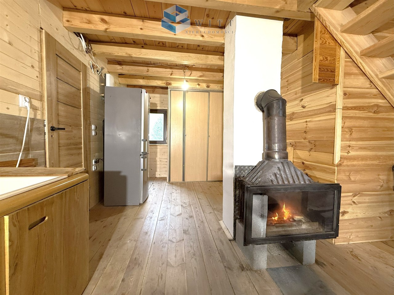 Wnętrze drewnianego domku z kuchnią, lodówką, kominkiem i drewnianymi schodami.