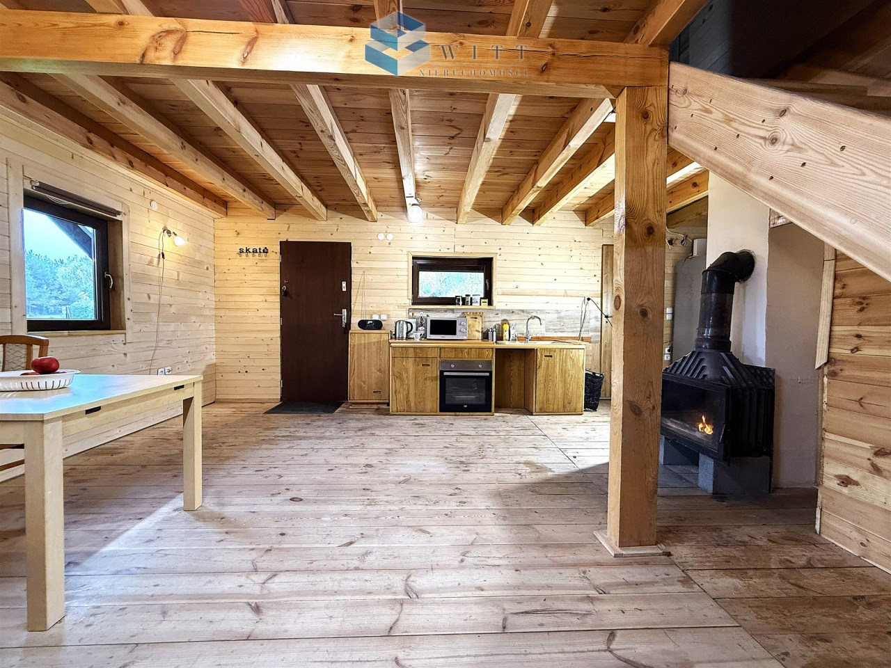 Wnętrze drewnianego domku z kuchnią, stołem i kominkiem. Ściany i sufit z naturalnego drewna, podłoga drewniana. Duże okna wpuszczające światło dzienne.