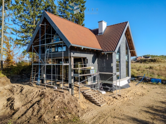 Nowoczesny dom jednorodzinny w trakcie budowy, z widocznymi rusztowaniami i częściowo wykończoną elewacją. W tle drzewa i otwarta przestrzeń.