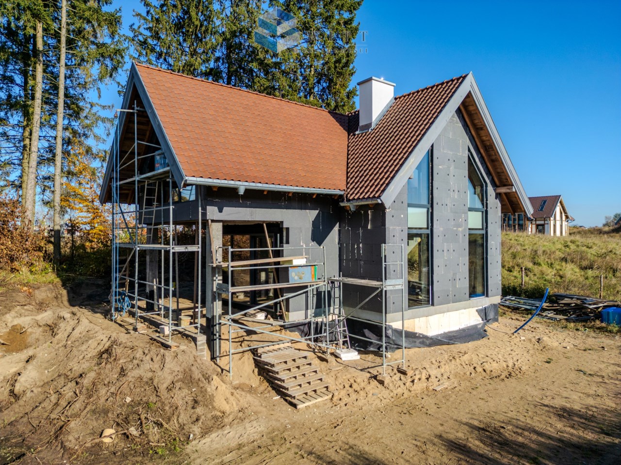 Nowoczesny dom jednorodzinny w trakcie budowy, z widoczną konstrukcją stalową i dużymi przeszkleniami. W tle drzewa i czyste niebo.