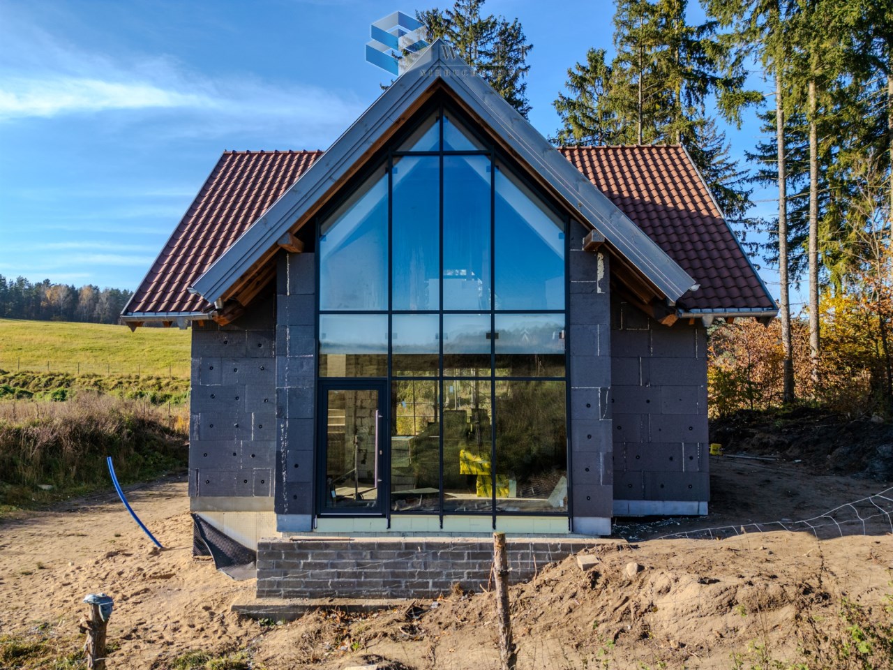Nowoczesny dom jednorodzinny w trakcie budowy, z dużymi przeszklonymi ścianami, dachówką oraz otoczeniem leśnym. W tle widoczna zielona łąka.