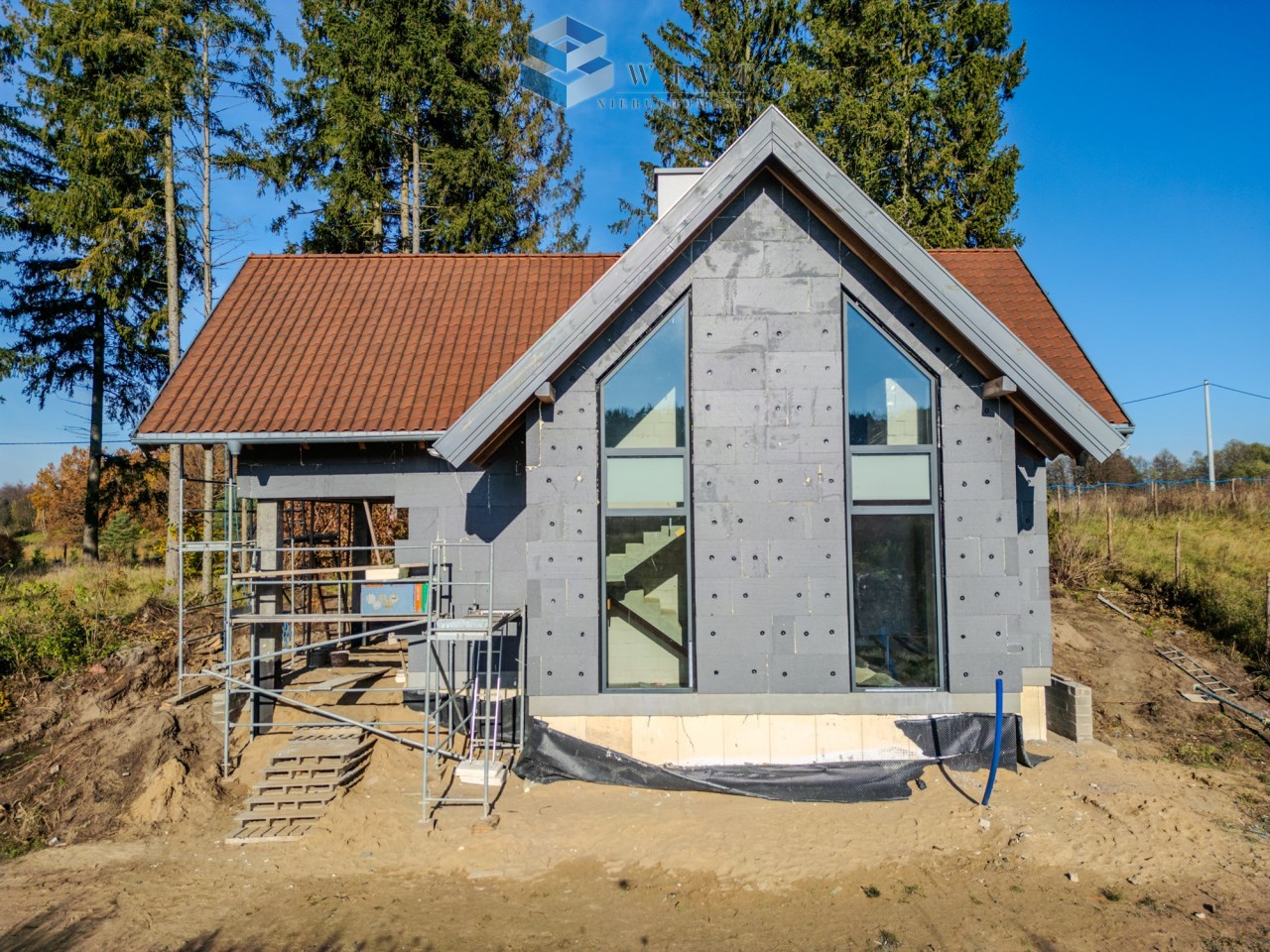 Zewnętrzny widok nowoczesnego domu w trakcie budowy, z dużymi oknami i dachem pokrytym dachówką, otoczony drzewami i placem budowy.