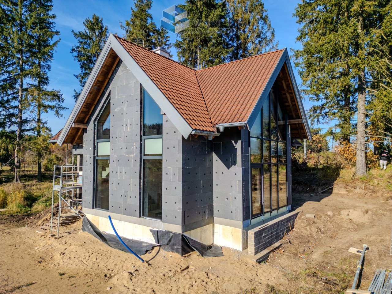 Nowoczesny dom jednorodzinny w trakcie budowy, z dużymi oknami, szarym ociepleniem i czerwoną dachówką, otoczony przez las. W tle widać rusztowanie oraz teren budowy.