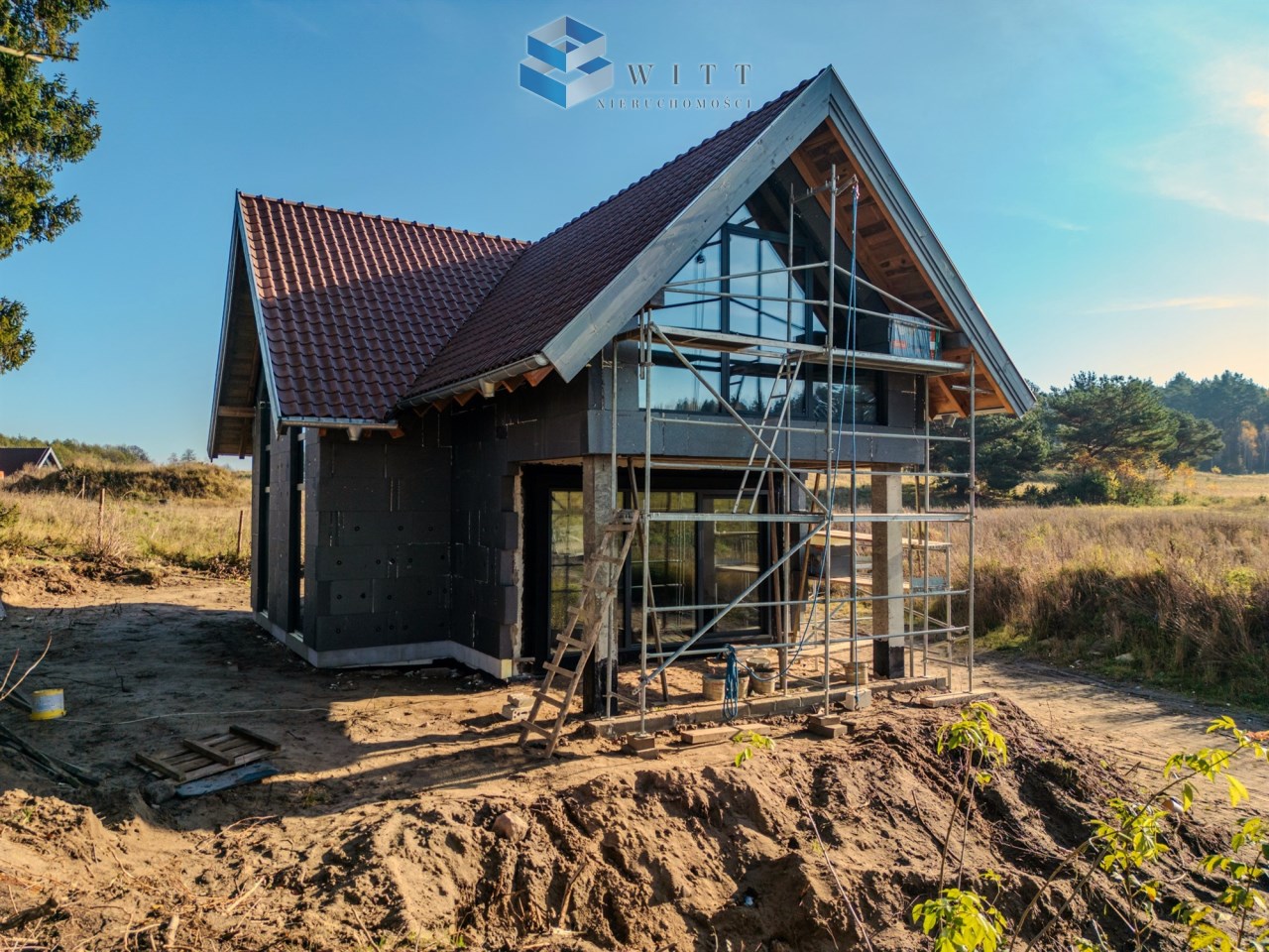 Nowoczesny dom w budowie z wielkimi oknami, otoczony rusztowaniem, na tle zielonego krajobrazu.
