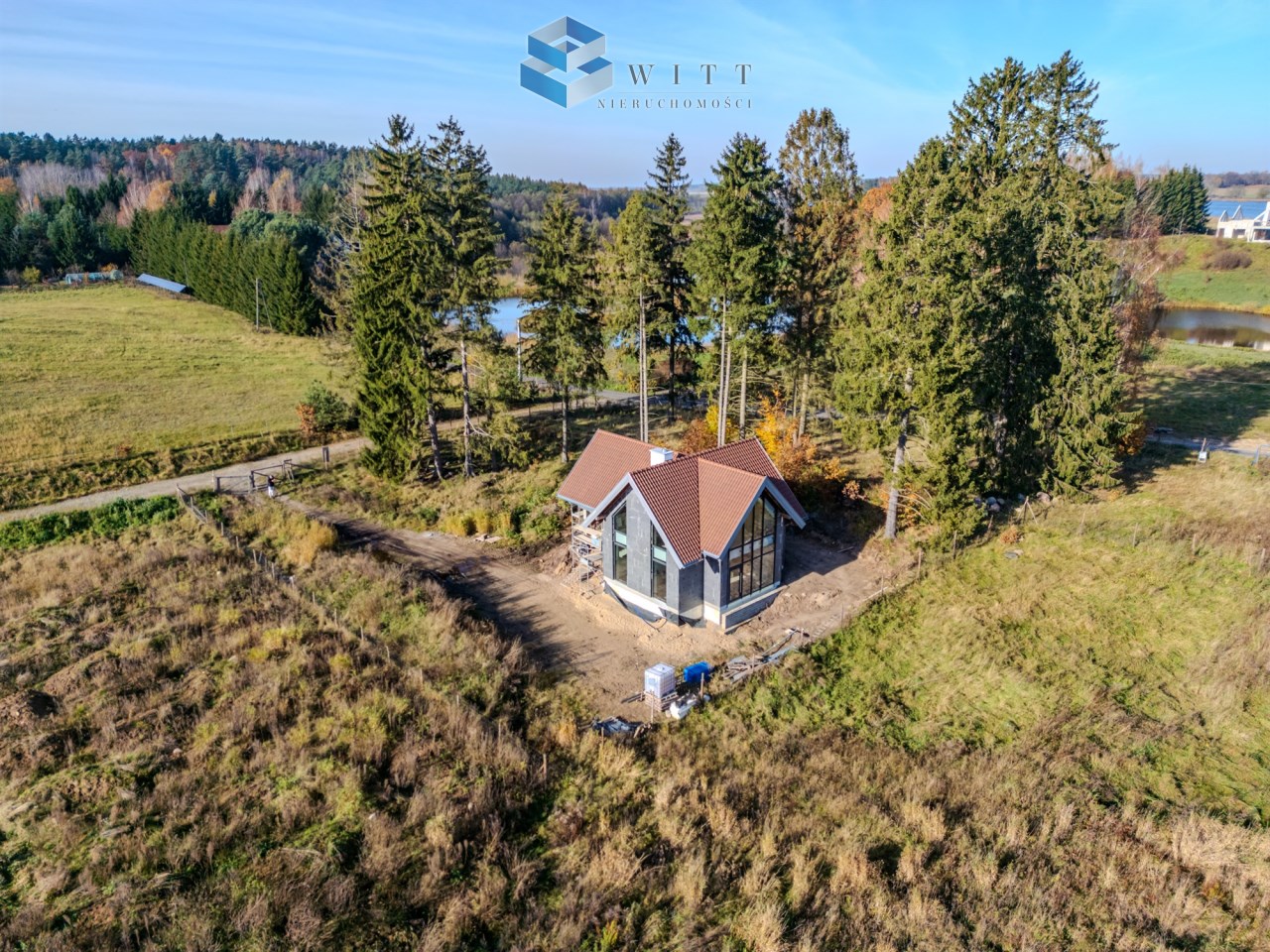 Nowoczesny dom z dużymi oknami, położony w malowniczej okolicy otoczonej lasem i wodą. Widok z góry, ukazujący tereny zielone i sąsiednie zabudowania.