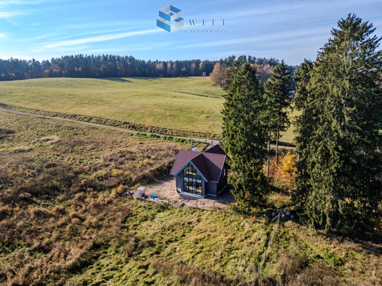Widok na nowoczesny dom położony w malowniczej okolicy, otoczony zielonymi łąkami i lasem.