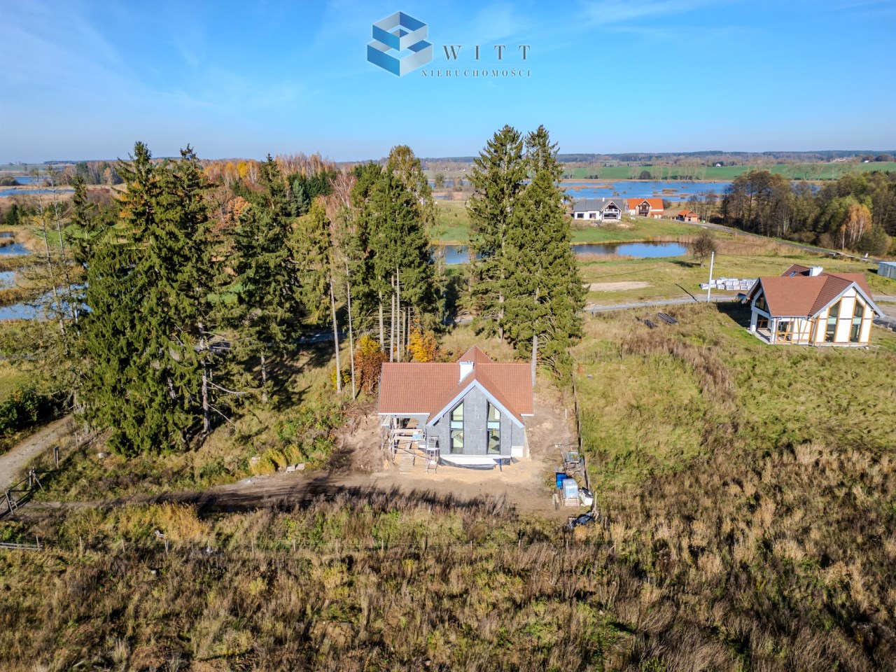 Widok z lotu ptaka na nowoczesny dom otoczony drzewami i zielenią, w tle jezioro i inne budynki. Idealne miejsce na wypoczynek i relaks w naturze.