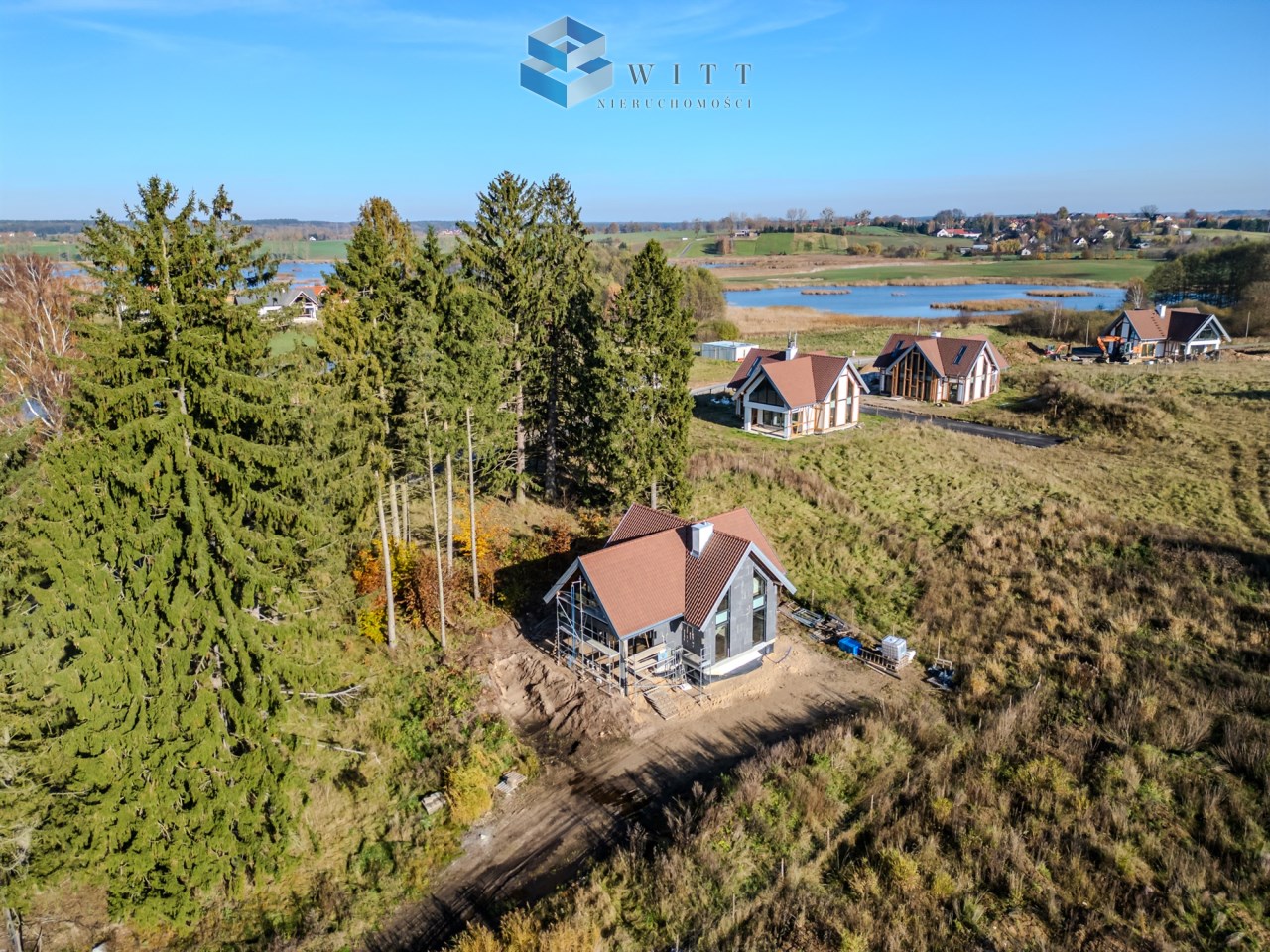 Widok na nowoczesne domy w otoczeniu zieleni, z drzewami w tle i wodą w oddali.