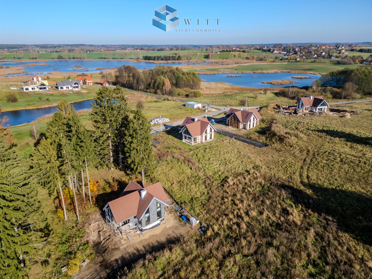 Widok z lotu ptaka na nowe domy w okolicy jeziora, otoczone zielenią i drzewami, w tle pola i lasy.