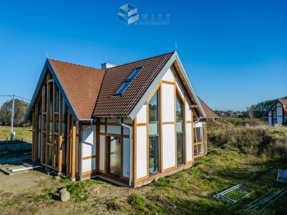 Nowoczesny dom jednorodzinny w budowie, z drewnianymi elementami i dużymi oknami, otoczony zielenią i widokiem na wiejskie tereny.